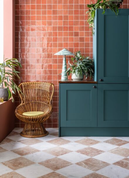 Zellige Coral Gloss Ceramic tiles on a kitchen wall, the backdrop of a navy set of cabinets and a wicker chair.